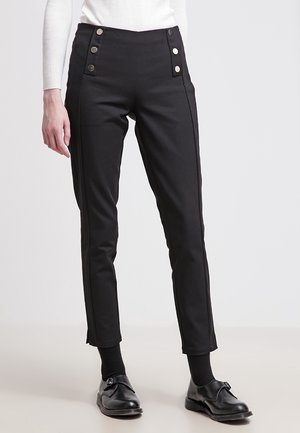 Femme portant un pantalon noir taille haute, coupe slim, avec des boutons décoratifs et des mocassins noirs, debout sur un fond clair.