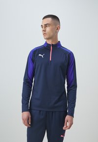 Tamsiai mėlynas sportinis ilgomis rankovėmis marškinėlis su violetiniais pečių elementais, užtrauktuko apykakle ir rožinės spalvos akcentais; ant krūtinės yra logotipas ir švelnaus audinio tekstūra.