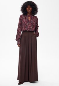 Vrouw met krullend haar in een patroon blouse met lange mouwen en een wijdvallende, bruine broek, staand met één hand in de zak.