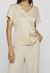 Beige, kortärmad blus med V-ringning, slätt tyg, avslappnad passform och diskret veck framtill, ihop med matchande byxor.