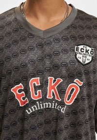 Harmaa lyhythihainen t-paita, jossa on toistuva kuvio, punainen ja valkoinen "ECKÖ unlimited" -teksti sekä embleemi vasemmassa rinnassa. V-aukko-muotoilu.