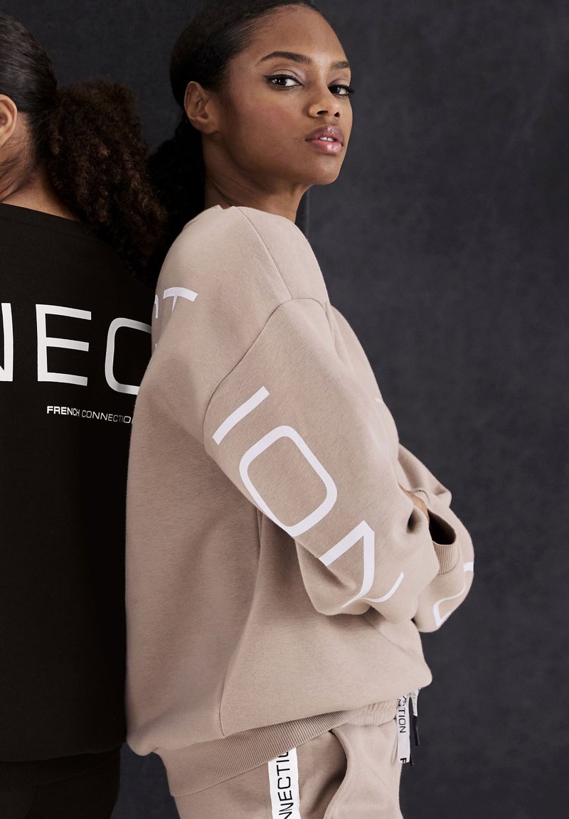 Beige sweatshirt met oversized pasvorm en witte tekst op de mouwen, gecombineerd met bijpassende broeken. Zwarte sweatshirt op de achtergrond weergegeven.