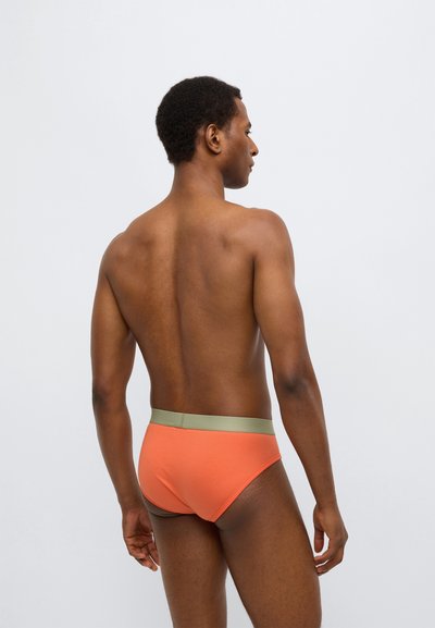 Herrunderbyxa i orange med ett grönt elastiskt midjeband, med en slät yta och åtsittande design, sedd bakifrån.