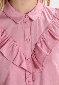 Camicia button-up rosa con una parte frontale arricciata, tessuto strutturato e piccoli motivi a pois. Presenta un colletto classico e bottoni coordinati.
