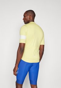Helles gelbes kurzärmliges Fahrradtrikot mit einem weißen Akzentband, kombiniert mit blauen Trägerhosen mit einer gepolsterten Einlage und Logodetails.