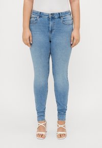 Kvinna iklädd ljusblå skinny jeans och vita remsandaler med hög klack, stående mot en neutral bakgrund.