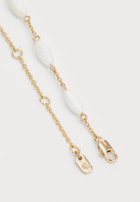 Zlatý reťazový náhrdelník s bielymi oválnymi korálkami, s karabínkovým zapínaním a gravírovanou visačkou s iniciálami, predvádzajúci jemný dizajn.