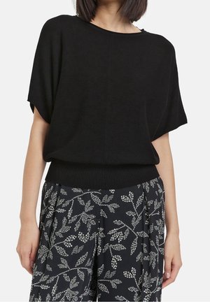 Vrouw draagt een losse zwarte top met korte mouwen, gecombineerd met zwarte wijde broek met een witte bladprint.