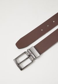 Curea din piele texturată de culoare maro, cu o cataramă rectangulară din metal argintiu ce prezintă un logo mic colorat și cinci găuri ajustabile.