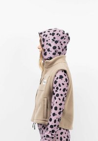 Beige Fleeceweste mit hohem Kragen und seitlichen Taschen, kombiniert mit einem pinken Hoodie mit weißen Punkten und schwarzen Flecken.