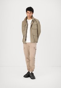 Jacket utilitar de culoare verde măsliniu, cu mai multe buzunare, asortat cu pantaloni cargo bej deschis, având un logo mic, și pantofi sport negri.