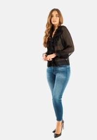 Blusa nera trasparente con dettagli a volant e maniche lunghe, abbinata a jeans blu aderenti e tacchi alti neri. Sfondo bianco.