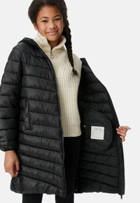 Giubbotto puffer nero con cuciture orizzontali, cappuccio, tasche frontali e chiusura con zip, indossato sopra un maglione lavorato a maglia crema. Etichetta interna visibile.
