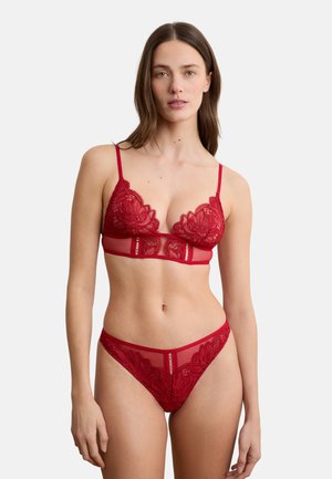 Frau trägt rotes Spitzen-BH und passende Spitzen-Pantys, steht vor einem schlichten hellen Hintergrund.