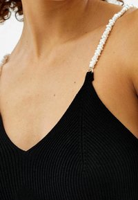 Närbild på en person som bär en svart ribbad topp med vita pärlade axelremmar, där övre bröst- och axelområdet visas.