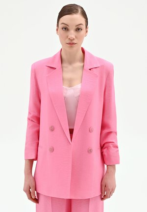 Femme portant un blazer rose vif croisé avec un pantalon assorti et un haut rose clair orné de dentelle sur un fond blanc uni.
