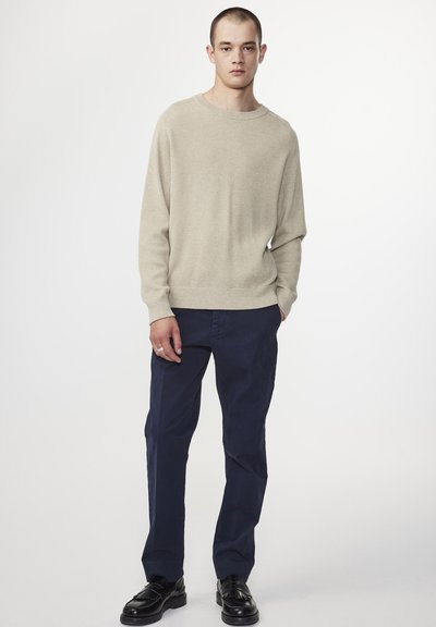 Beiger RIP-Knit Pulli mit Rundhalsausschnitt, kombiniert mit dunkelblauen Hosen und schwarzen Schuhen, vor einem weißen Hintergrund dargestellt.