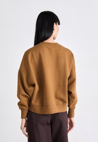 Felpa oversized marrone con scollo rotondo e spalle scese. Tessuto morbido, polsini e orlo a costine. Indossata con pantaloni marroni scuri.