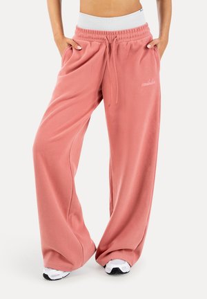 Różowe szerokie spodnie dresowe z białym elastycznym paskiem w talii, bocznymi kieszeniami oraz białe trampki, noszone przez osobę stojącą z rękami w kieszeniach.