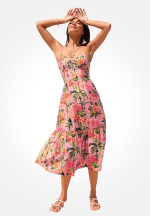 Frau in einem rosa floralen Midikleid mit dünnen Trägern und Sandalen, die mit erhobenen Händen über dem Kopf und geschlossenen Augen posiert.