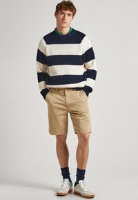 Suéter de punto con amplias rayas en azul marino y crema, combinado con pantalones cortos beige. Zapatillas blancas y calcetines azul marino completan el atuendo.