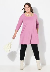 Rosa Tunika-Kleid mit drei Viertelärmeln, kombiniert mit schwarzen Leggings und weißen Ankle Boots. Accessoires: eine weiße Handtasche und eine auffällige Halskette.