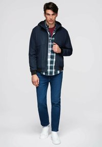 Námořnická bunda s kapucí, nošena přes kostkovanou košili, v kombinaci s modrými džíny a bílými teniskami. Hladký materiál; přiléhavý a neformální design.