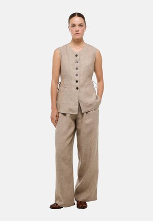 Femme debout portant un gilet beige sans manches boutonné et un pantalon large assorti avec des chaussures tissées marron, sur un fond blanc uni.