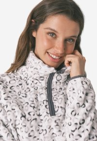 Løveprint fleecejakke i hvid og grå med højt krave, frontlynlås og blød tekstur. Har grå pynt langs lynlåsen.