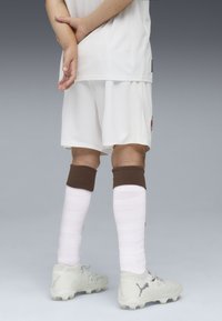 Vit fotbollströja och shorts, vita strumpor med bruna manschetter, ljusgrå fotbollsskor med texturerad ovandel och dobbar, modell stående med armarna i kors.