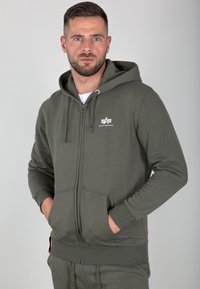 Sweat à capuche zippé en vert olive, avec un logo blanc sur la poitrine, des poignets côtelés et des poches avant pour un design fonctionnel.