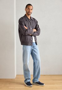 Gray cotton jacket with button closure, white t-shirt, light blue jeans, and navy sneakers with textured white soles. Standing pose on wood floor.