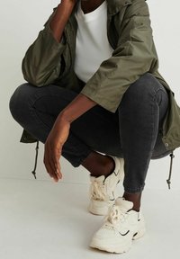 Veste légère vert olive portée sur une chemise blanche texturée, associée à un jean skinny noir et des baskets beiges avec des accents noirs.