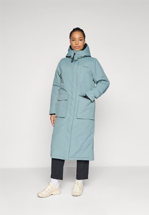 Vrouw in een lange lichtblauwe winterjas met capuchon en grote zakken aan de voorkant, zwarte broek, witte sokken en beige sneakers, staande tegen een effen achtergrond.