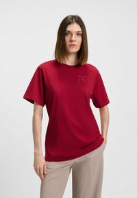 Rød kortærmet t-shirt lavet af glat stof med rund halsudskæring og en subtil logo detaljeret på venstre bryst. Kombineret med beige bukser.