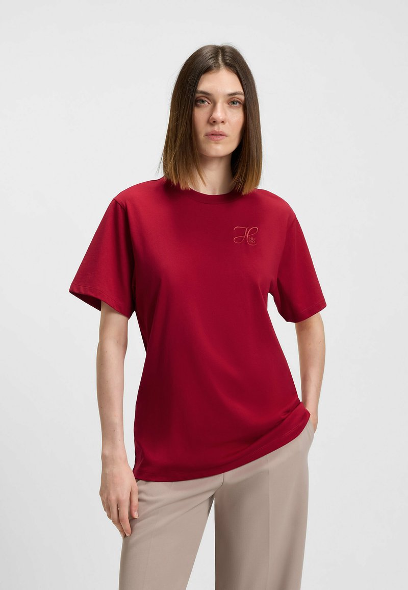 Rød kortærmet t-shirt lavet af glat stof med rund halsudskæring og en subtil logo detaljeret på venstre bryst. Kombineret med beige bukser.