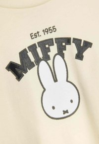 Crème-kleurig shirt met een grafisch ontwerp van Miffy met een witte konijn en zwarte omtrekken. "MIFFY" in vette zwart bovenaan gedrukt. Tekst "Opgericht 1955" is ook inbegrepen.