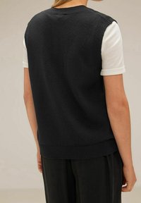 Black sleeveless sweater with ribbed hem, worn over a white shirt. Soft texture and relaxed fit, showing a clean, minimalist design.