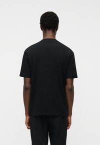 Άνδρας με κοντά σγουρά μαλλιά που φοράει μαύρο απλό κοντομάνικο πουκάμισο και μαύρο παντελόνι, στέκεται γυρισμένος με την πλάτη σε λευκό φόντο.