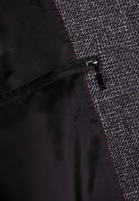 Sort lynlås lommeforing ved siden af tekstureret grå stof, med røde stitching detaljer og "HUGO" logo synligt på det sorte materiale.