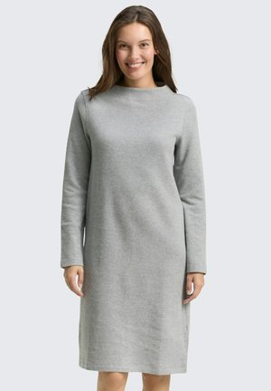 Lächelnde Frau trägt ein knielanges, langärmeliges, hellgraues Strickkleid vor einfachem hellem Hintergrund.