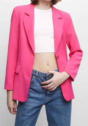 Vrouw draagt een felroze blazer, wit crop top en blauwe spijkerbroek met één hand op haar taille tegen een effen achtergrond.