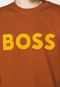 Sudadera marrón con un atrevido texto amarillo "BOSS" en el pecho. La tela parece suave con un cuello redondo y puños acanalados.