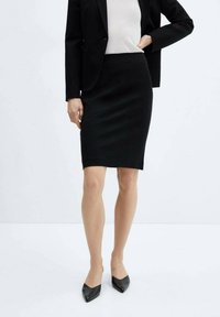 Jupe crayon noire, longueur au genou, en tissu lisse avec une coupe ajustée. Portée avec un haut de couleur claire et des escarpins noirs à bout pointu.