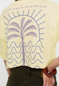 T-shirt jaune avec un motif de palmier violet, des vagues bleues et des rayons de soleil. Le texte au-dessus indique : « Vous ne pouvez pas arrêter la vague, mais vous pouvez apprendre à surfer. »