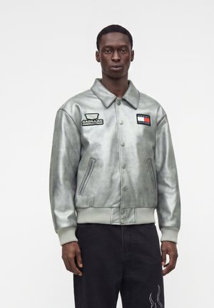 Homme portant une veste argentée boutonnée avec les logos de l'équipe Cadillac de Formule 1 et Tommy Hilfiger, et un pantalon noir avec des motifs de flammes sur la cuisse.
