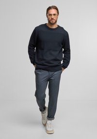 Un uomo con barba e occhiali cammina in avanti indossando un maglione scuro e texture, pantaloni grigi e sneakers bianche su uno sfondo semplice.