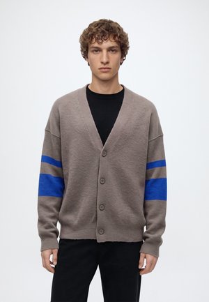 Jeune homme aux cheveux bouclés portant un cardigan taupe avec des rayures bleues sur les manches, par-dessus une chemise noire, debout devant un fond blanc uni.