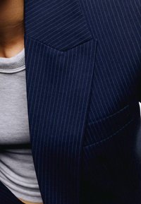 Chaqueta de traje a rayas marinas con solapa muesca y un bolsillo en el pecho, combinada con una camiseta de cuello redondo gris claro debajo.