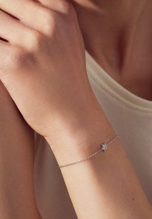 Silverarmband med en stjärnformad berlock som har en klar ädelsten, fäst på ett fint kedja, som vilar på ett handled mot en neutral bakgrund.